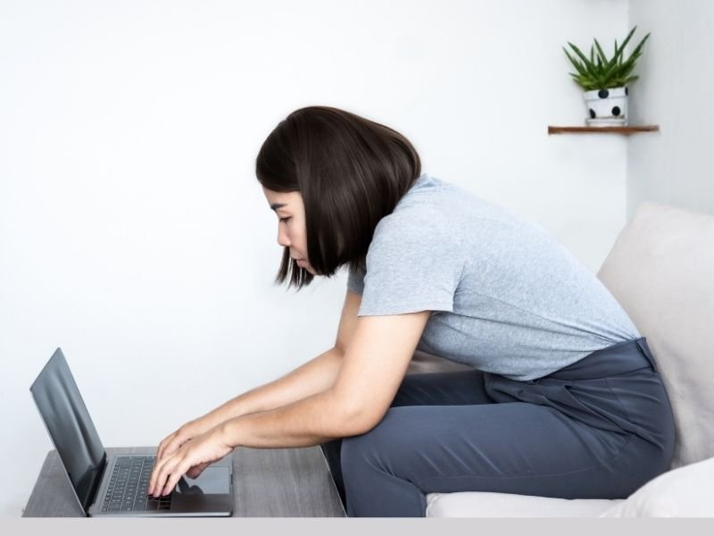 Imagem mostrando uma mulher escrevendo no computador com postura incorreta, prejudicando uma boa postura